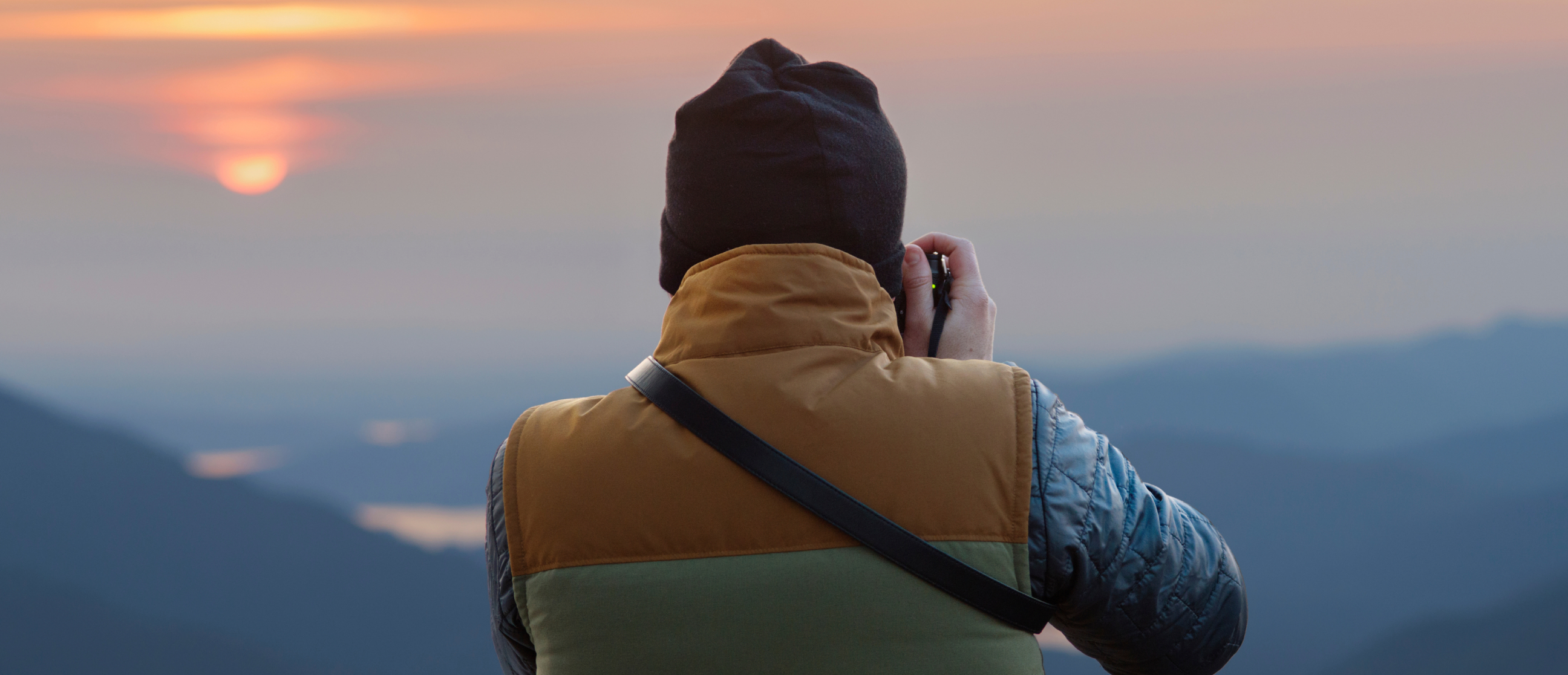 Looking over the shoulder of a person taking a photo of a mountain sunset.