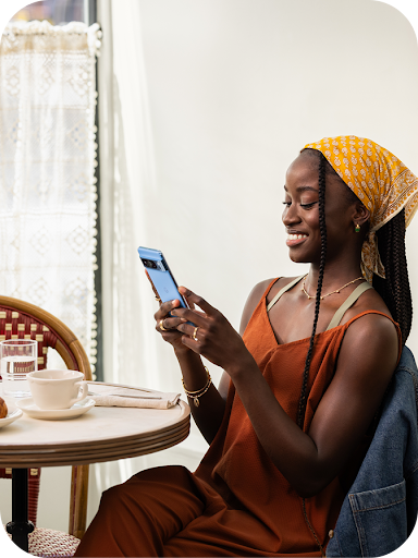 A person with long braids, wearing a yellow headscarf and a rust-colored dress, sits at a table and smiles while using a smartphone. The setting appears to be a bright cafe or restaurant.