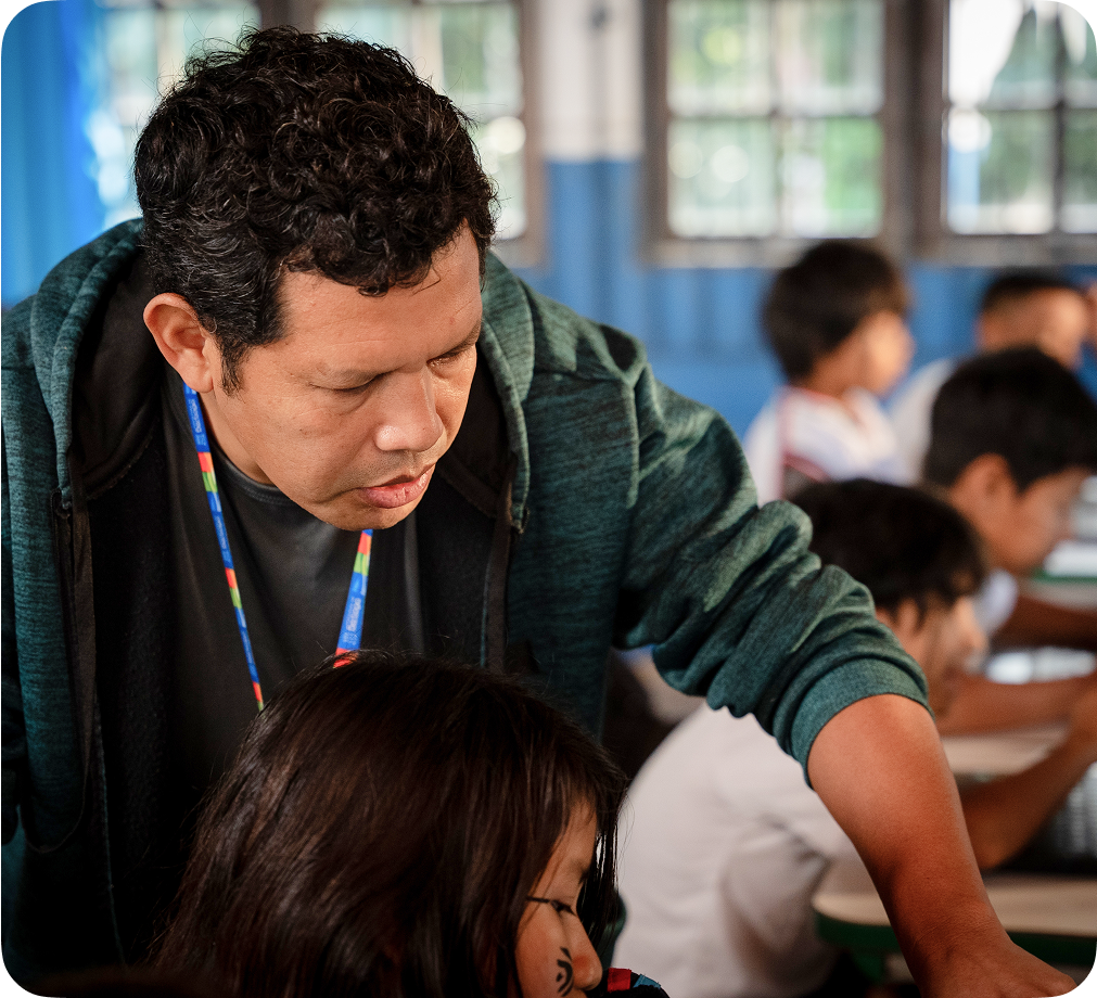 Un profesor en un aula inclinándose para ayudar a un estudiante que está trabajando en un dispositivo Chromebook.