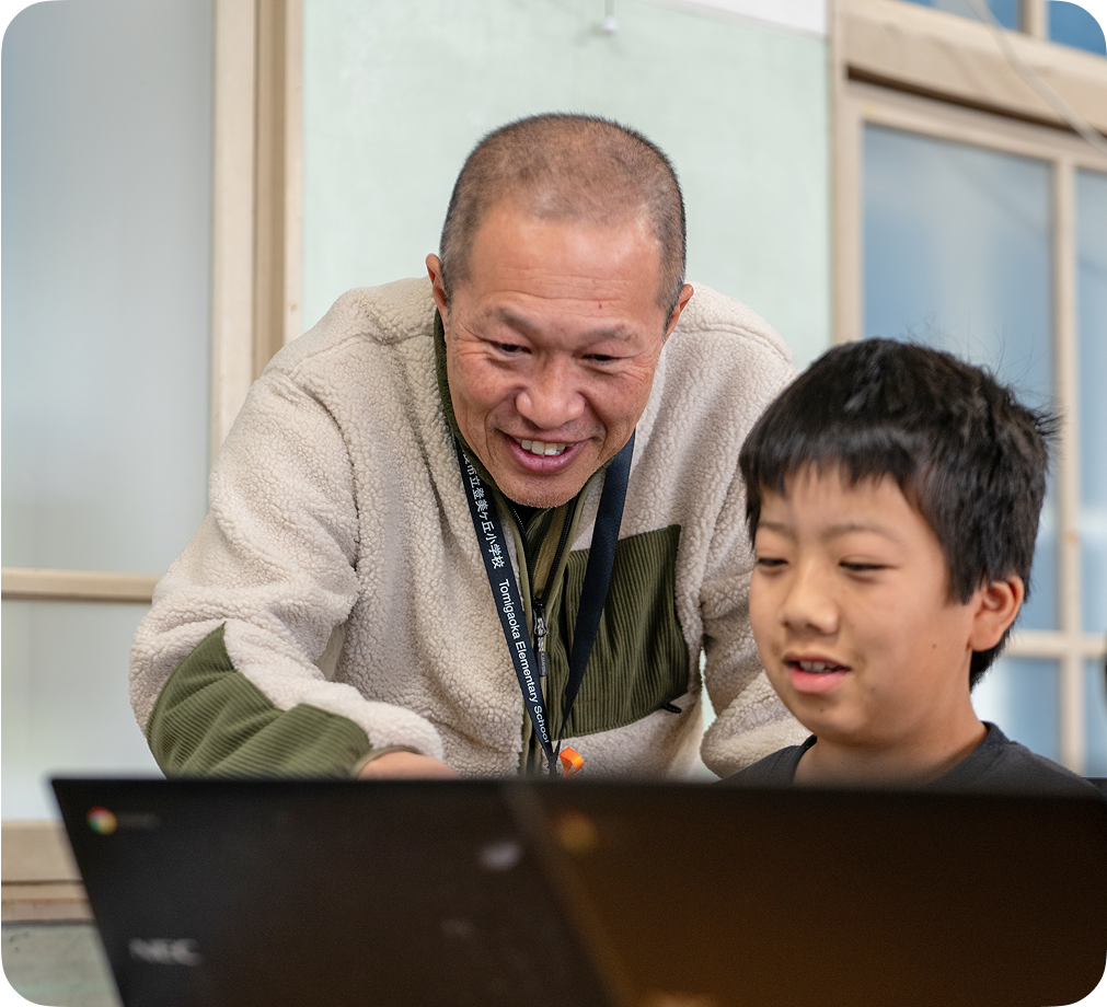 教室で Chromebook デバイスを使って一緒に作業している笑顔の教師と児童。